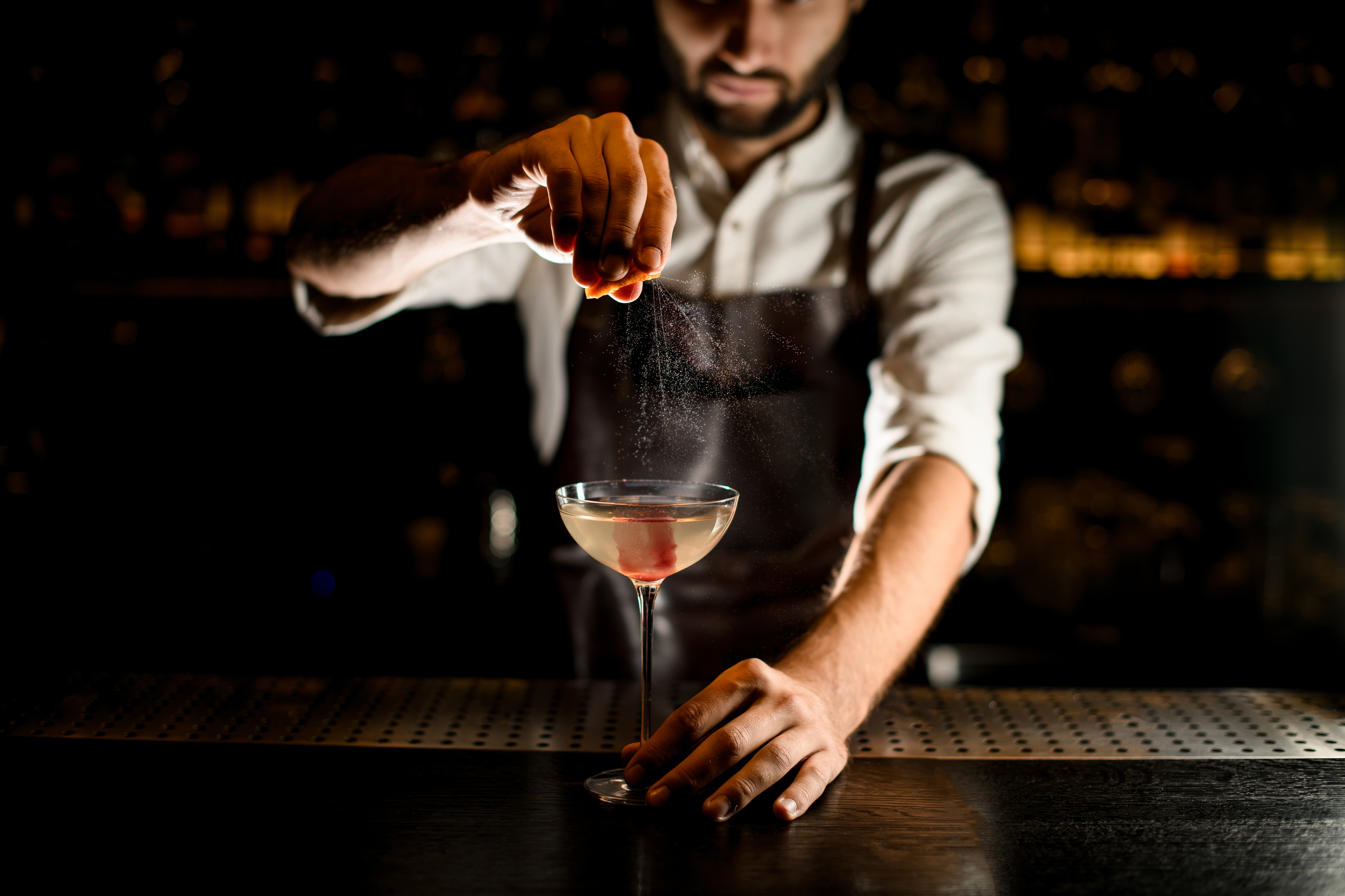 Professional male bartender serving a cocktail in the glass decorated with pink ice cube adding a lemon juice on the bar counter